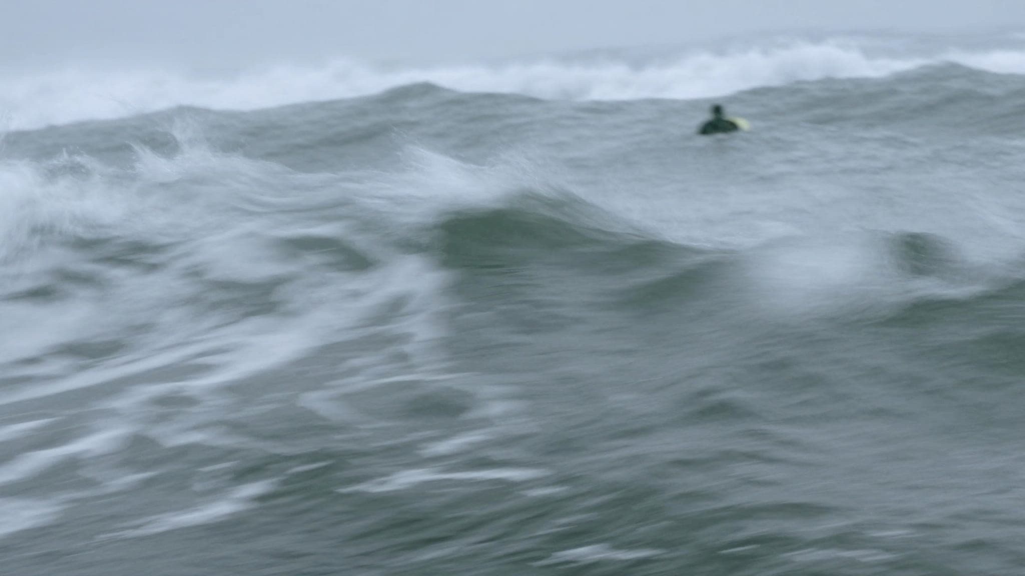 Line-up mosso visto dall'acqua: onda grigio-blu disordinata che sale verso l'obiettivo, un surfista lontano seduto di spalle sulla tavola.
